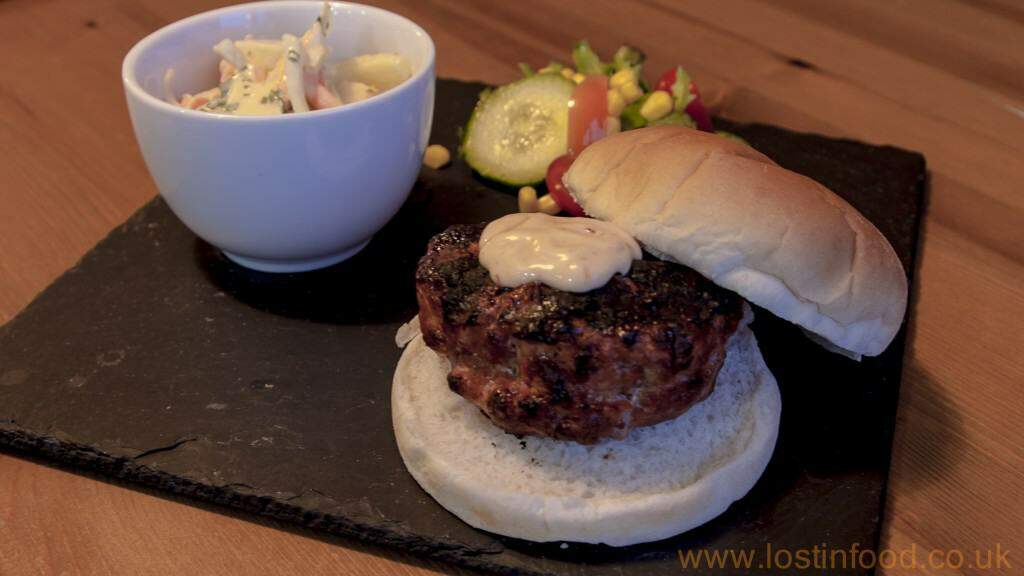 A burger bun filled with pork burger, salad and topped with mayonnaise on a black slate plate with a bowl of coleslaw.