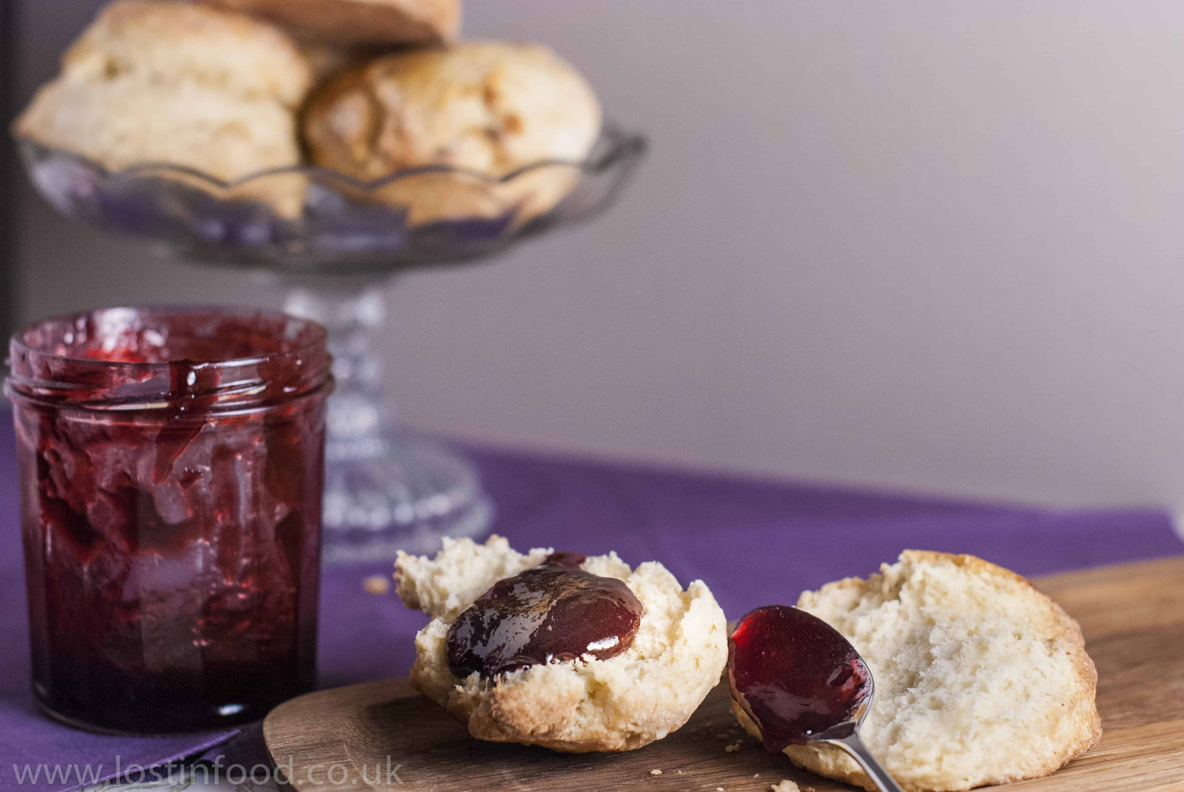 A glass cake stand of buttermilk scones and an open buttermilk scone topped with fresh homemade jam and the jar of jam open to the side.