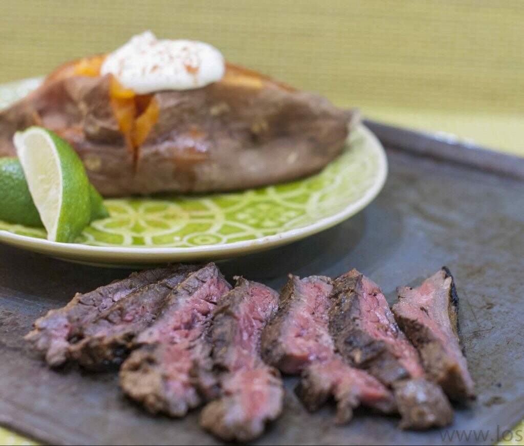 A black tray with rare cooked and sliced skirt steak and a green plate with a baked sweet potato topped with sour cream and a wedge of lime.