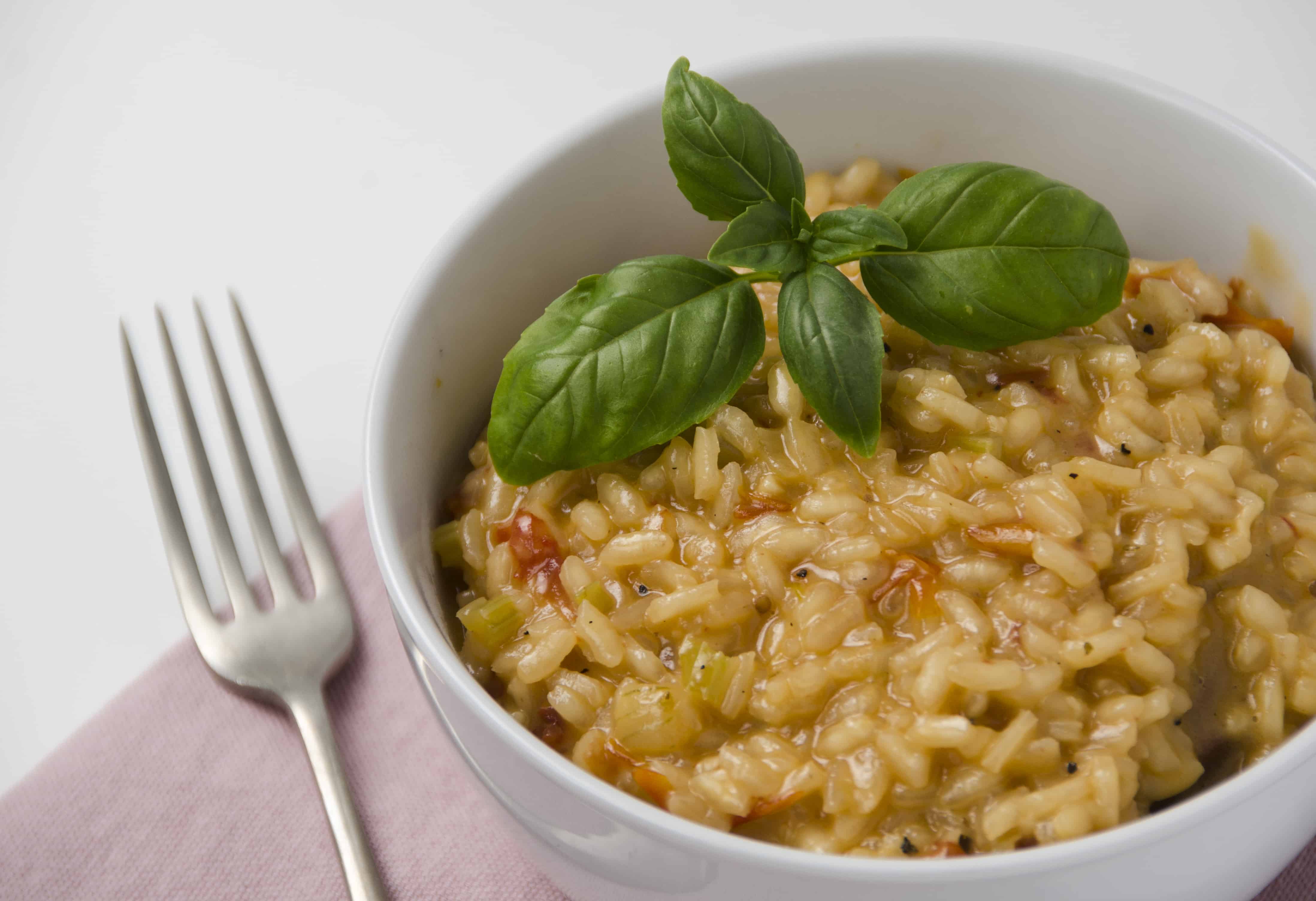 A bowl of tomato based risotto garnished with basil and served in a white bowl on a pink napkin with a fork to the side.