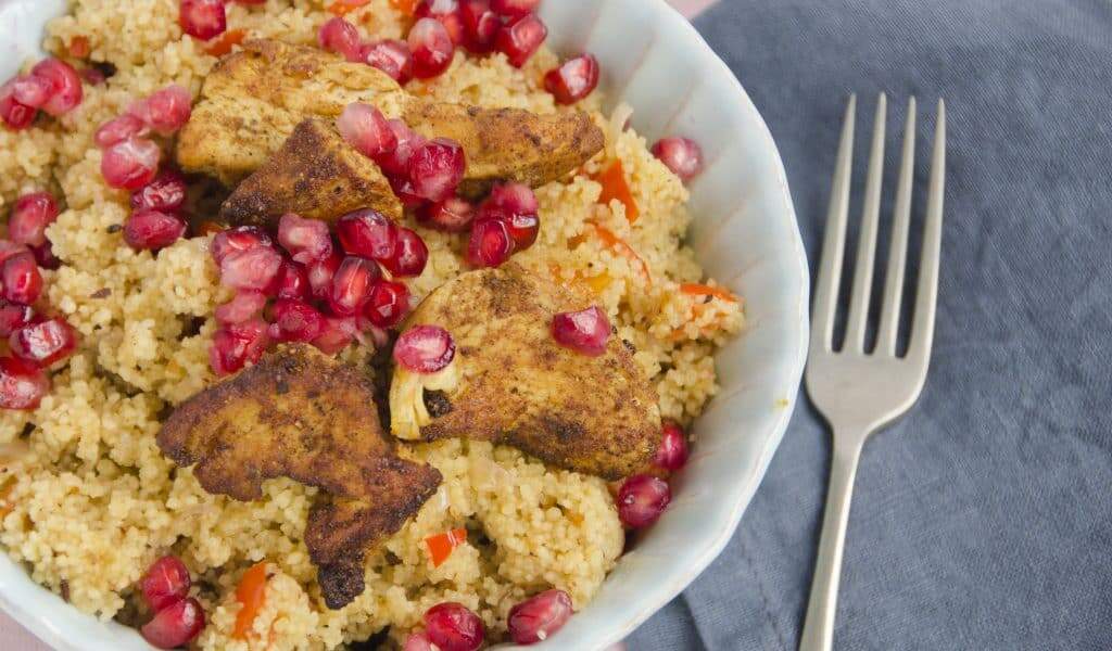 A large bowl of spiced couscous topped with marinated and grilled chicken breast and garnished with pomegranate seeds.