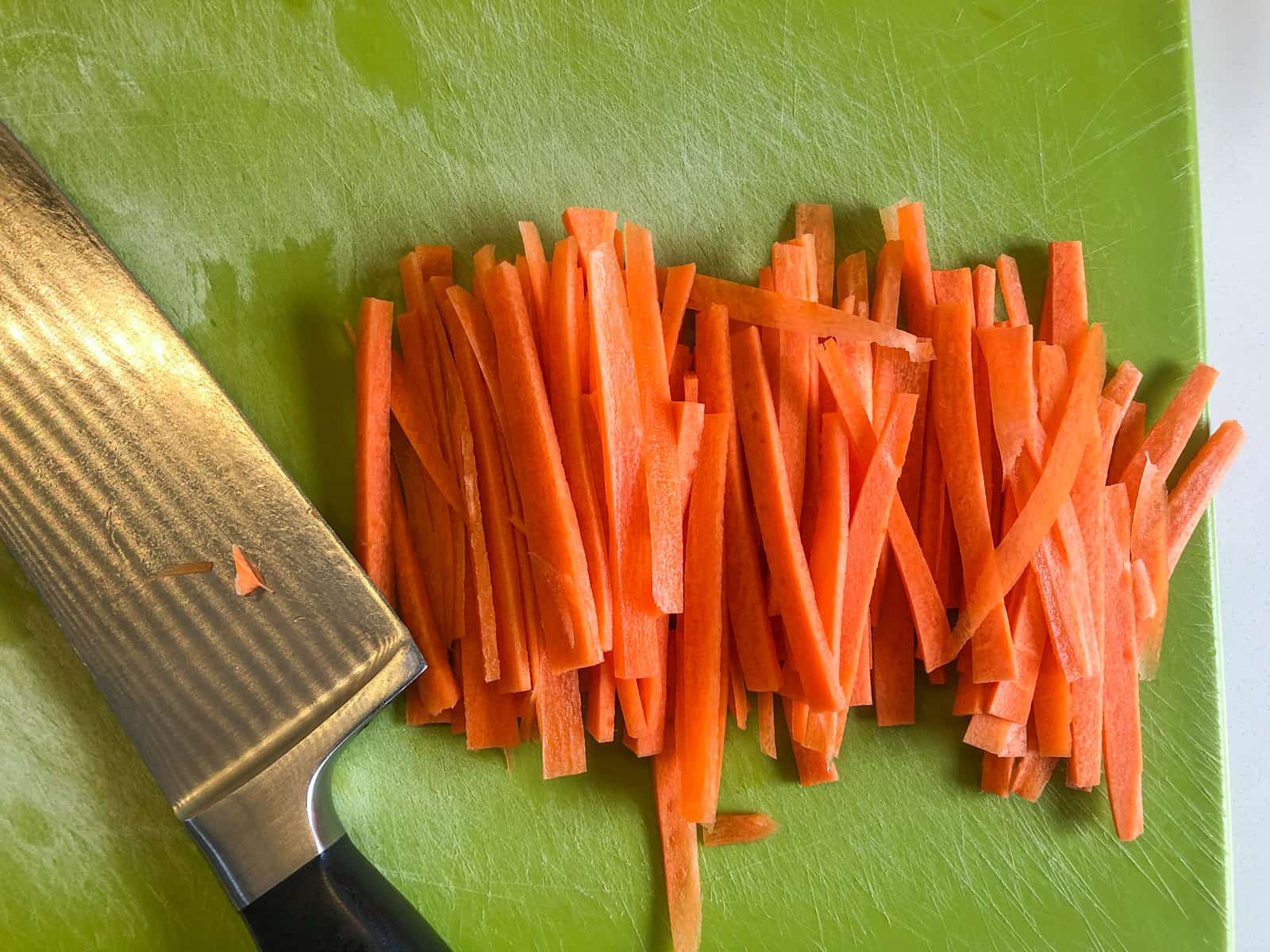 Julienned carrots on a green board with a sharp knife to the side.