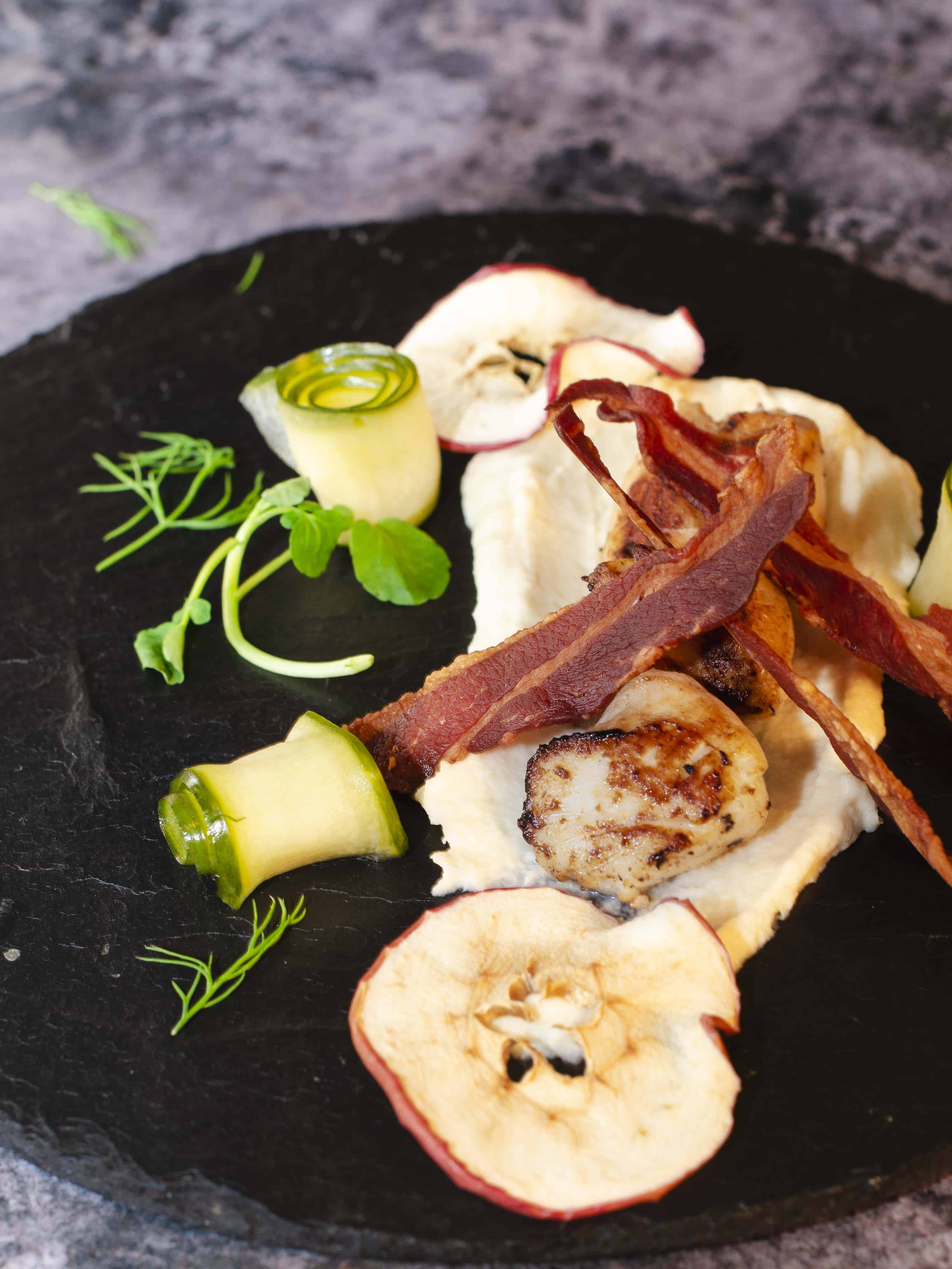 scallops on celeriac puree. dried apple on a black slate garnished with bacon shards