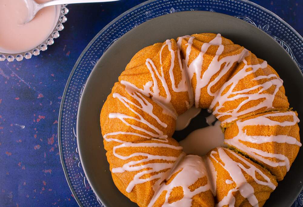 a top view of a blood orange drizzle cake with a blood orange drizzle icing on a glass plate layered with a grey plate, on a dark blue backdrop and a glass dish of icing in the background