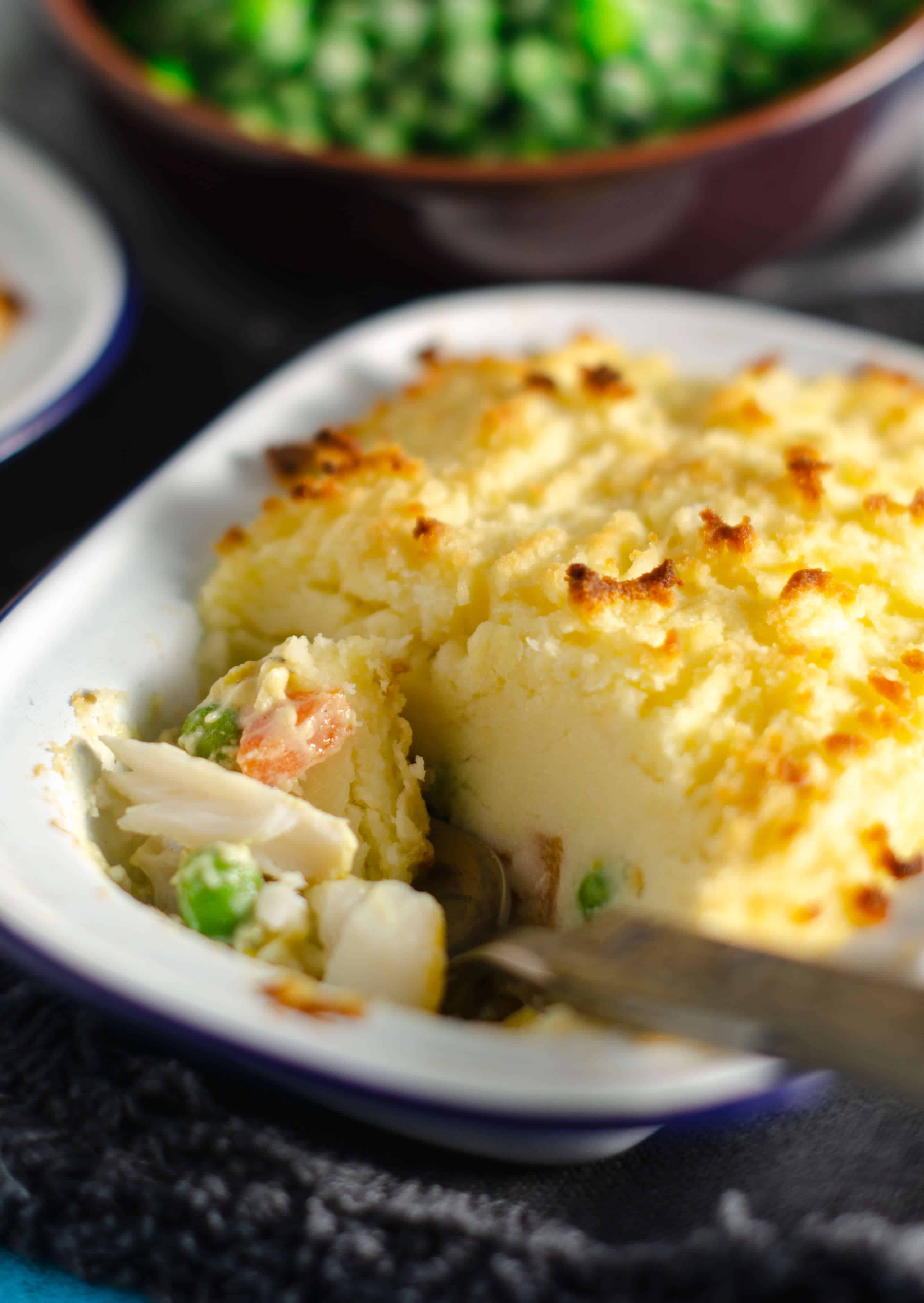 A closeup image of an individual fish pie with a spoon tucking in and a bowl of peas just barely visible in the background.