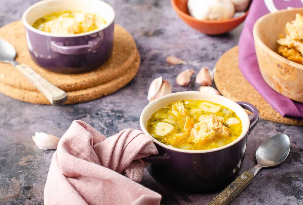 two bowls of clear garlic soup with sourdough croutons in le crueset purple dishes and pink napkins and rustic spoons