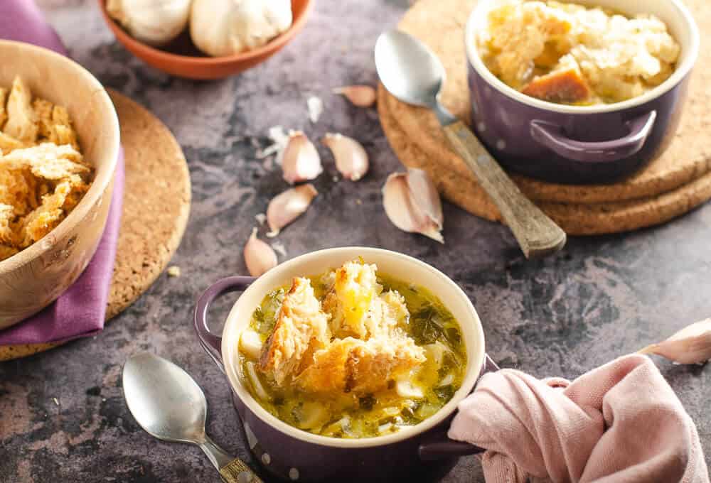 A warming soothing bowl of garlic broth soup in purple le crueset bowls on a dark marbled backdrop, scattered garlic cloves and matching pink linen napkins