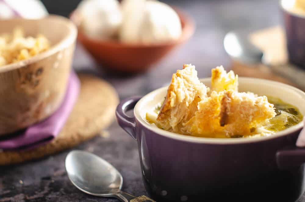 Sourdough croutons on top of a bowl of clear garlic broth soup in purple le crueset bowls