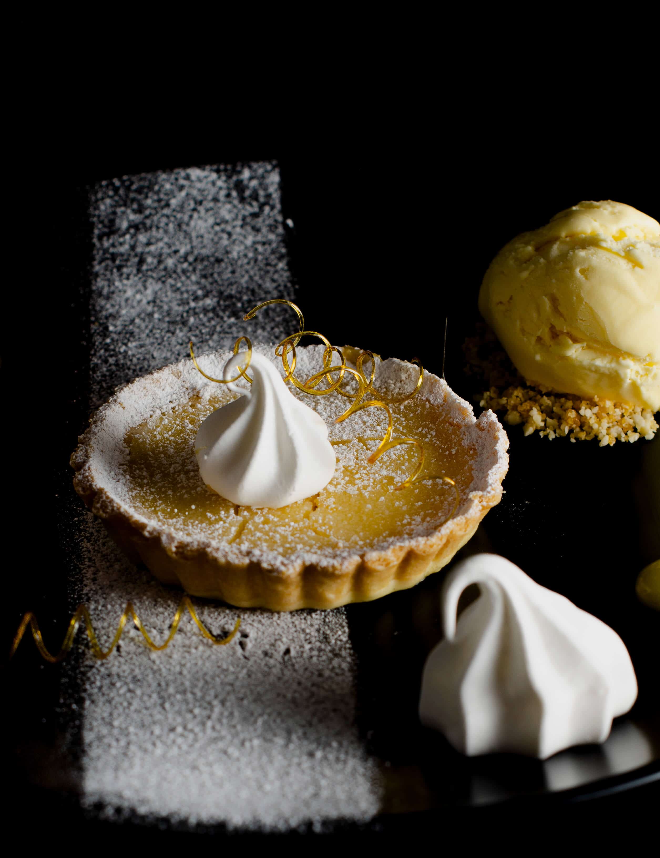 lemon tart with limoncello ice cream and sugar spirals on a black plate in a dark background