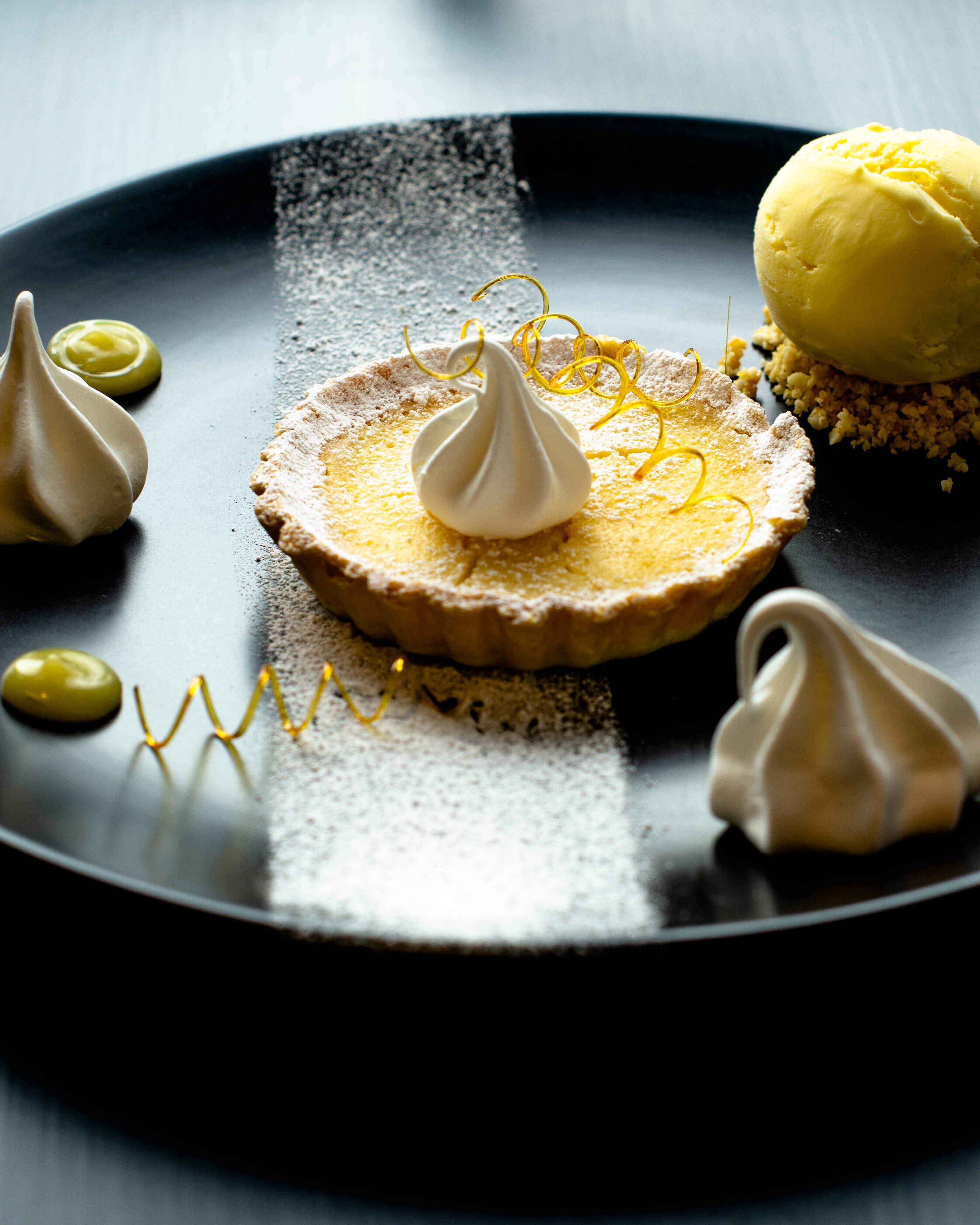 lemon tart with mini meringues and sugar spirals with a limoncello ice cream on the side, on a black plate and dark background with the light catching from the back