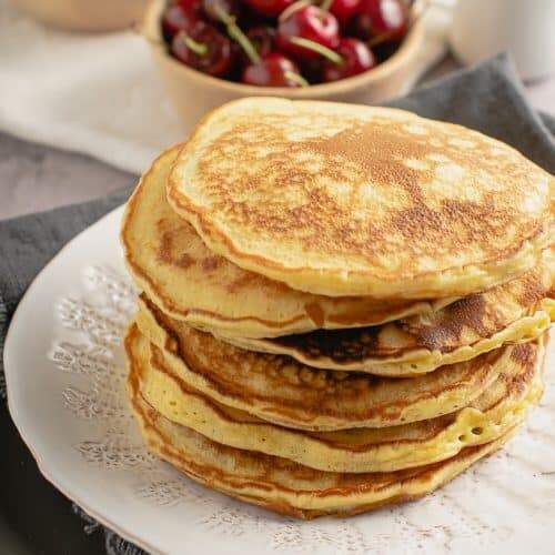 a stack of scotch pancakes on a white plate with cherries and blueberries in the background and a small jug pf maple syrup