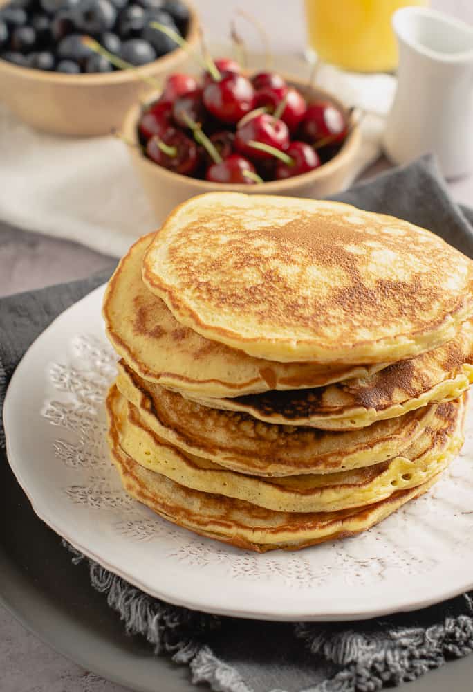 a stack of scotch pancakes on a white plate with cherries and blueberries in the background and a small jug pf maple syrup