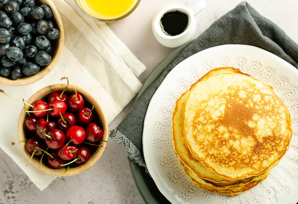 a flat lay of a breakfast table with scotch pancakes, orange juice, maple syrup, cherries, blueberries on a marble effect table