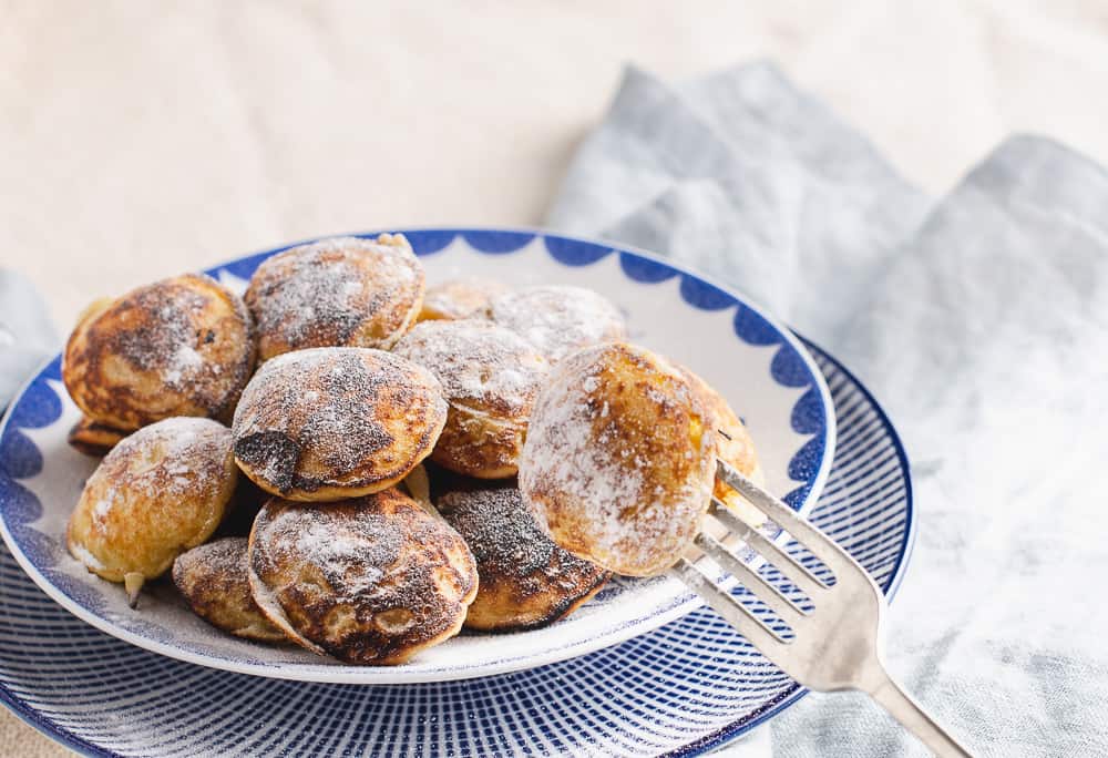 an inviting plate of dutch mini pancakes (pofferjes) topped with sugar and a blue background with a fork holding a pancake ready to eat