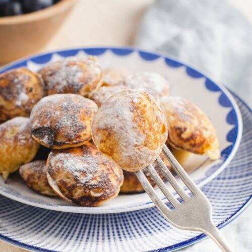 a blue and white backdrop with a plate of dutch mini pancakes (pofferjes) and blueberries in the background topped with sugar