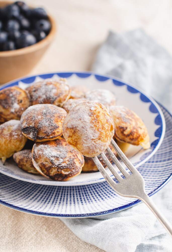 a blue and white backdrop with a plate of dutch mini pancakes (pofferjes) and blueberries in the background topped with sugar