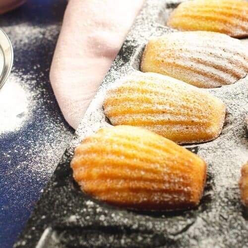 Close up of Madeleines in a black baking tray dusted with icing sugar