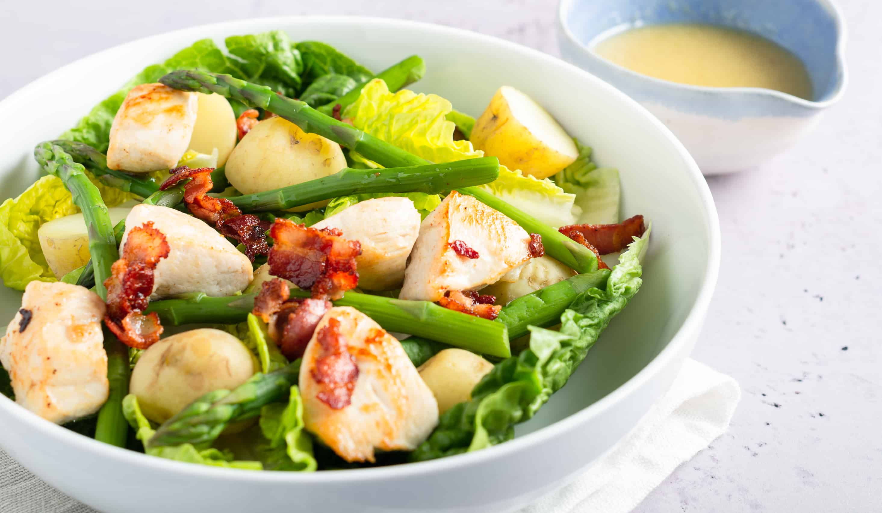Green salad in a white bowl with cubed chicken, fresh cooked warm new pototoes, crispy bacon and a pouring dish of salad dressing.