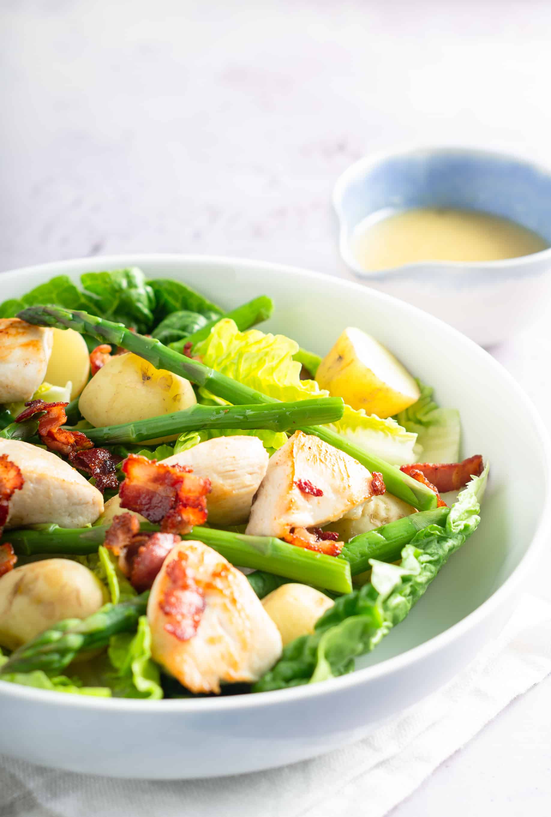 A spring bowl of salad with new potatoes, asparagus, crispy bacon and diced chicken in a white bowel and a blue bowl filled with salad dressing