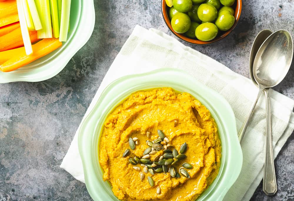 bowl of carrot hummus in a green bowl with vegetable sticks and olives in the back