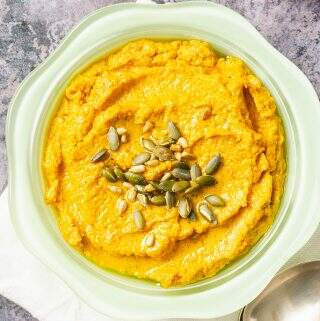 close up of a bowl of carrot hummus with spoons and a bowl of olives just peaking in the corner