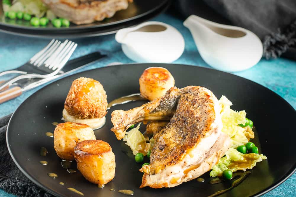 Guinea Fowl with pan fried greens, fondant potato and chicken leg bonbon with a chicken jus on a black plate in a table setting