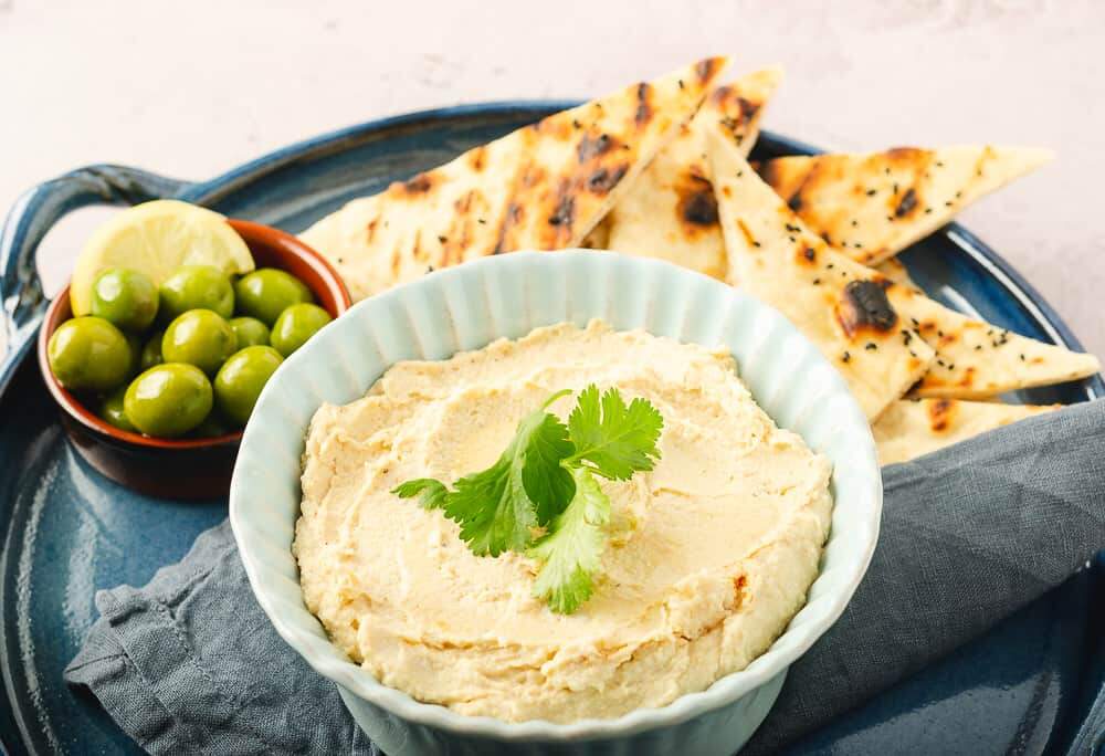 a big bowl of chickpea hummus in a blue bowl on a blue napkin and tray with a bunch of triangle shaped flatbreads in the back