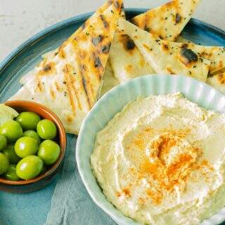 a close up of a bowl of chickpea hummus in a blue bowl sat on a blue napkin and a blue ceramic tray with flatbreads and a bowl of olives