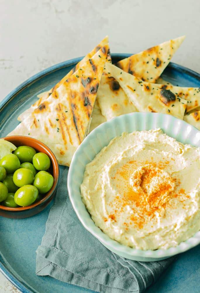 a close up of a bowl of chickpea hummus in a blue bowl sat on a blue napkin and a blue ceramic tray with flatbreads and a bowl of olives