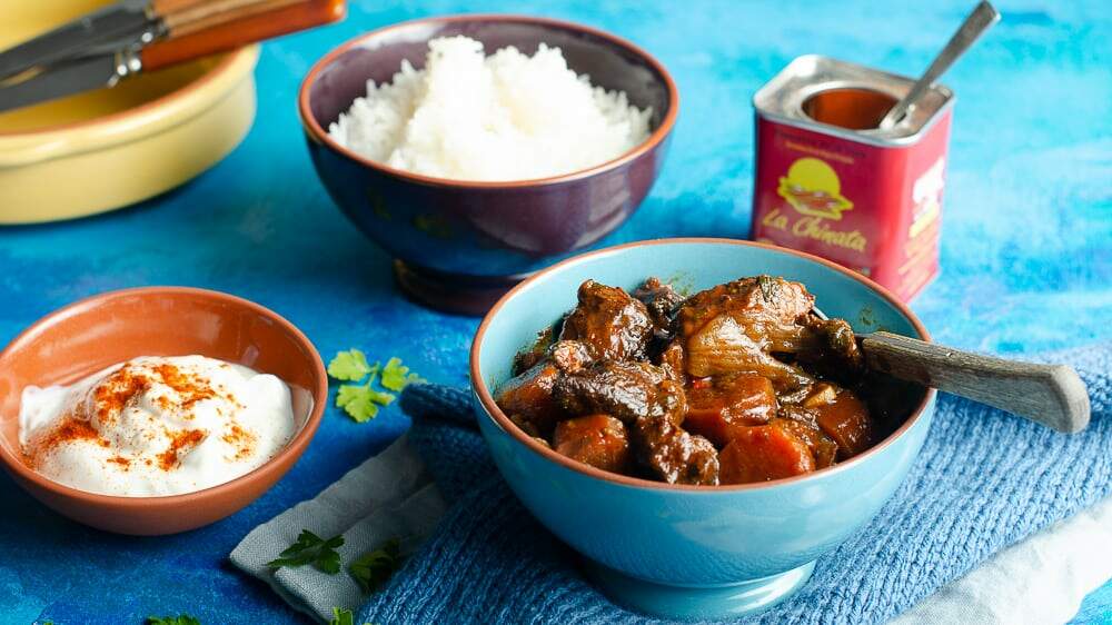 smoked paprika beef casserole served with a bowl of rice on a blue backdrop with blue napkins and a tin of smoked paprika in the back
