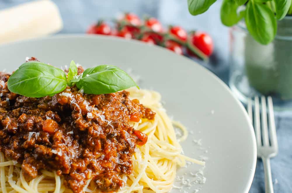 A plate of Spaghetti Bolognese with vine tomatoes, a basil plant and parmesan cheese topped with basil spring