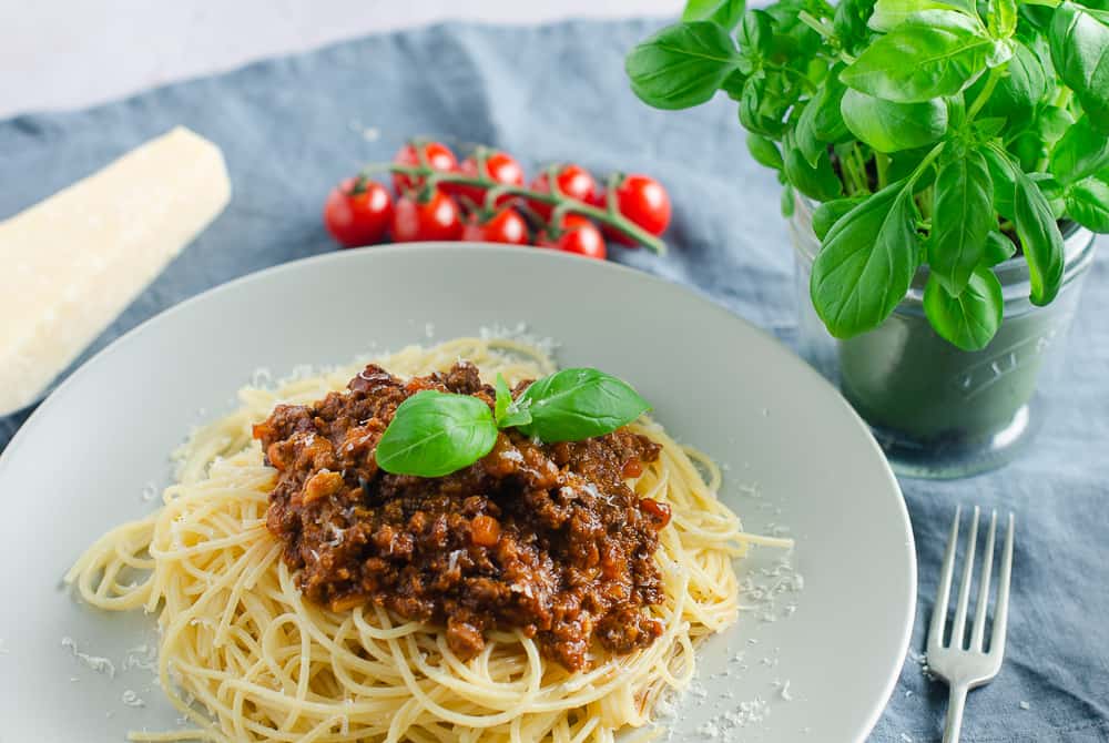 a large plate of spaghetti bolognese on a blue background with a basil plant, vine tomatoes and a block of parmesan cheese in the back.