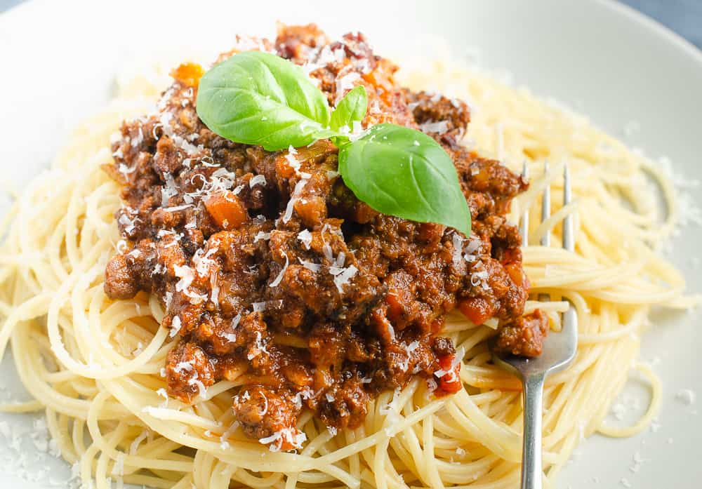 a grey plate with a large pile of spaghetti bolognese with a fork ready to eat on a light grey plate topped with a couple of basil leafs
