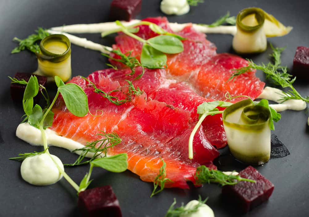 A closeup view of a black matt plate serving of sliced beetroot cured salmon on top of a horseradish cream with cubes of beetroot, slices of pickled cucumber rolled and avocado dil cream served around the plate and finished with fresh dill and pea shoots.