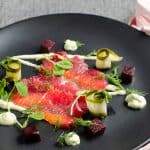 A stunning black plate with slices of beetroot cured salmon, cubed beetroot, a drizzle of horseradish cream, rolled slices of pickled cucumber, dollops of avocado dill cream and topped with fresh sprigs of dill and pea shoots.