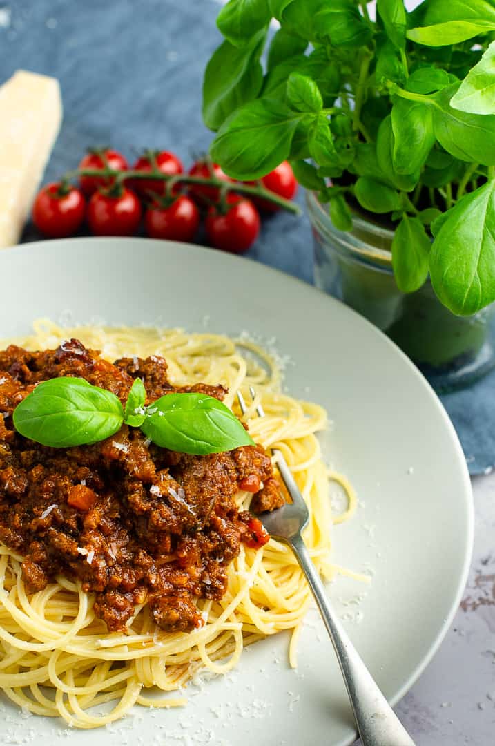 A pale grey plate filled with spaghetti bolognese topped with basil leaves and fresh tomatoes and basil in the back.