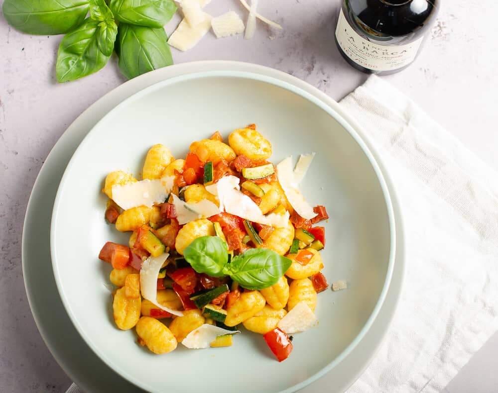 View from the top of a bowl of gnocchi tossed with diced chorizo, fresh tomatoes, diced courgettes and topped with shavings of parmesan and fresh basil with tomatoes and olive oil in the background.