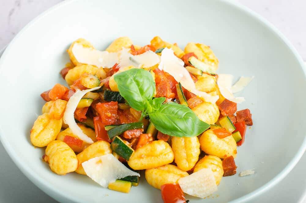 A closeup of a bowl of gnocchi tossed with chopped fresh tomatoes, diced chorizo and courgettes and finished with shavings of parmesan and basil leaves.