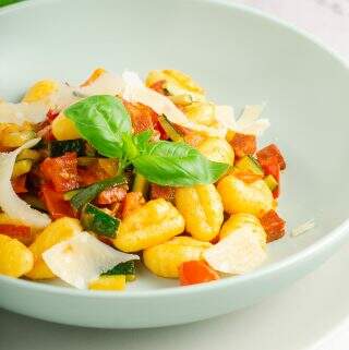 A vertical image of a bowl of gnocchi with fresh tomatoes, chorizo, courgettes and garlic with added shavings of parmesan and basil.