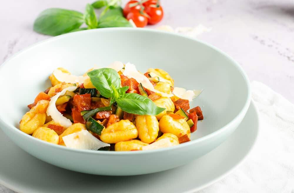 A pale green bowl on a plate filled with gnocchi tossed in fresh tomatoes, garlic, diced courgettes and fresh tomatoes and then topped with fresh basil leaves and shavings of parmesan.