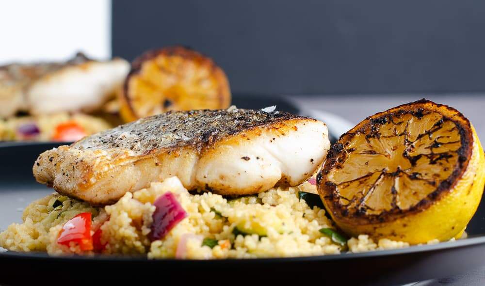 Looking straight ahead at an image of pan fried hake on vegetable and lemon couscous with a grilled lemon half on the side.