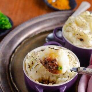 2 individual indian spiced cottage pies in purple le creuset casseroles on a tarnished metal tray with a spoonful ready to eat, steamed broccoli in the back with curry powder in a black dish all on an old mahogany table.