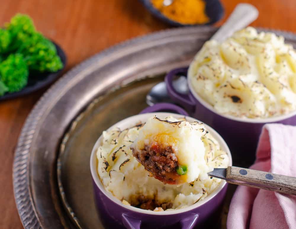 2 individual indian spiced cottage pies in purple le creuset casseroles on a tarnished metal tray with a spoonful ready to eat, steamed broccoli in the back with curry powder in a black dish all on an old mahogany table.