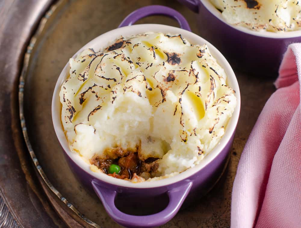 an individual indian spiced cottage pies in purple le creuset casseroles on a tarnished metal tray with a spoonful removed to see the inside filled with curry spiced mince, carrots and peas.