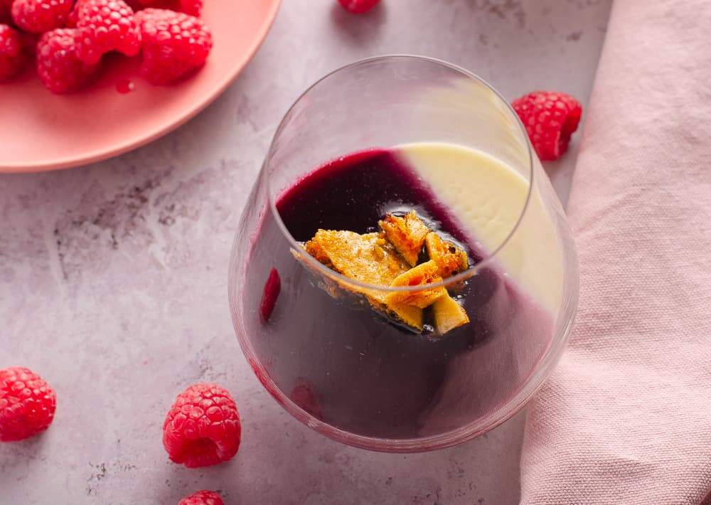Peering into a glass of fresh lemon pannacotta with fresh raspberry jelly and honeycomb on top, with a plate of fresh raspberries and a pink napkin.