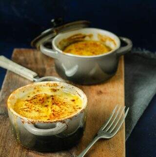 Two individual casserole pots of a creamy vegetable bake topped with melted cheese and served on a dark wooden board.