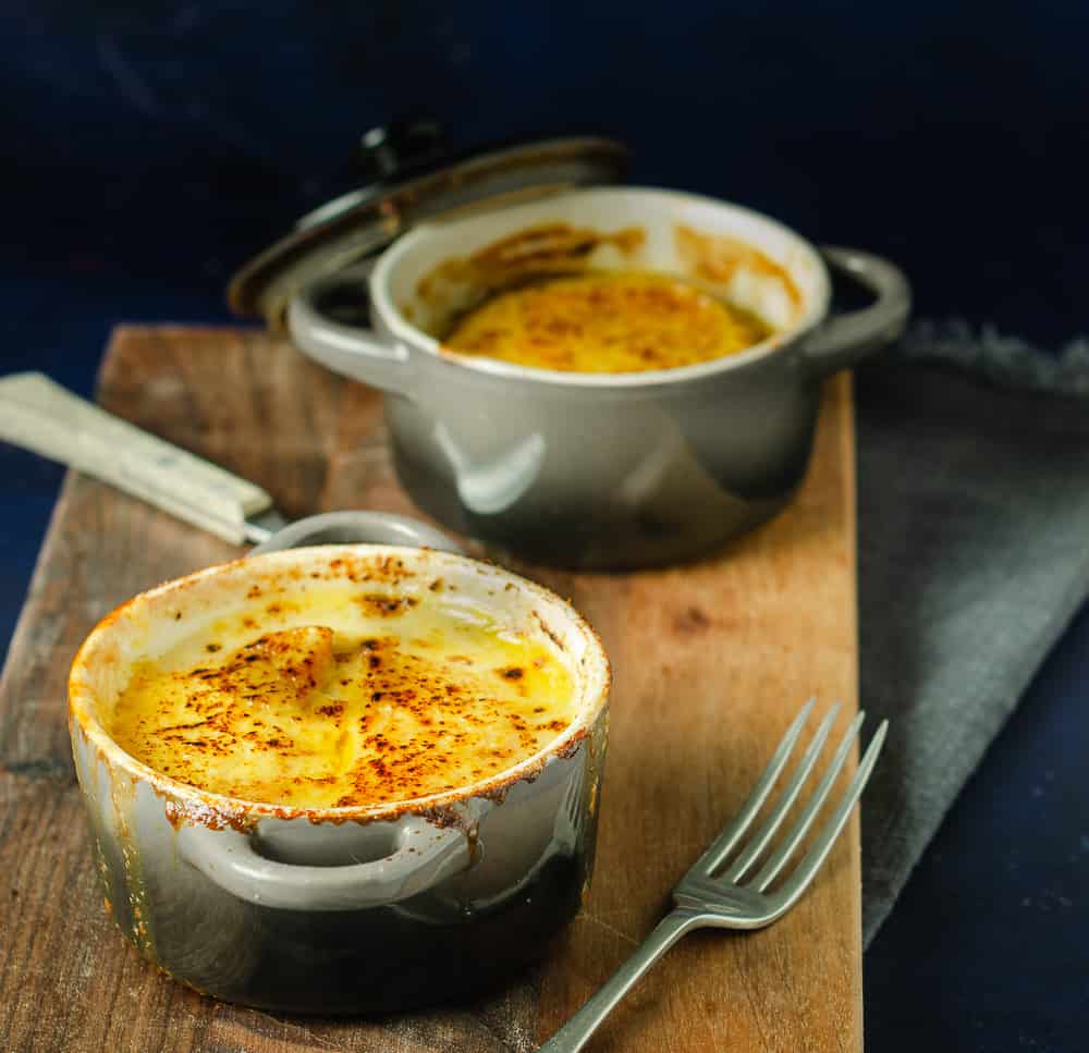 Two individual casserole pots of a creamy vegetable bake topped with melted cheese and served on a dark wooden board.