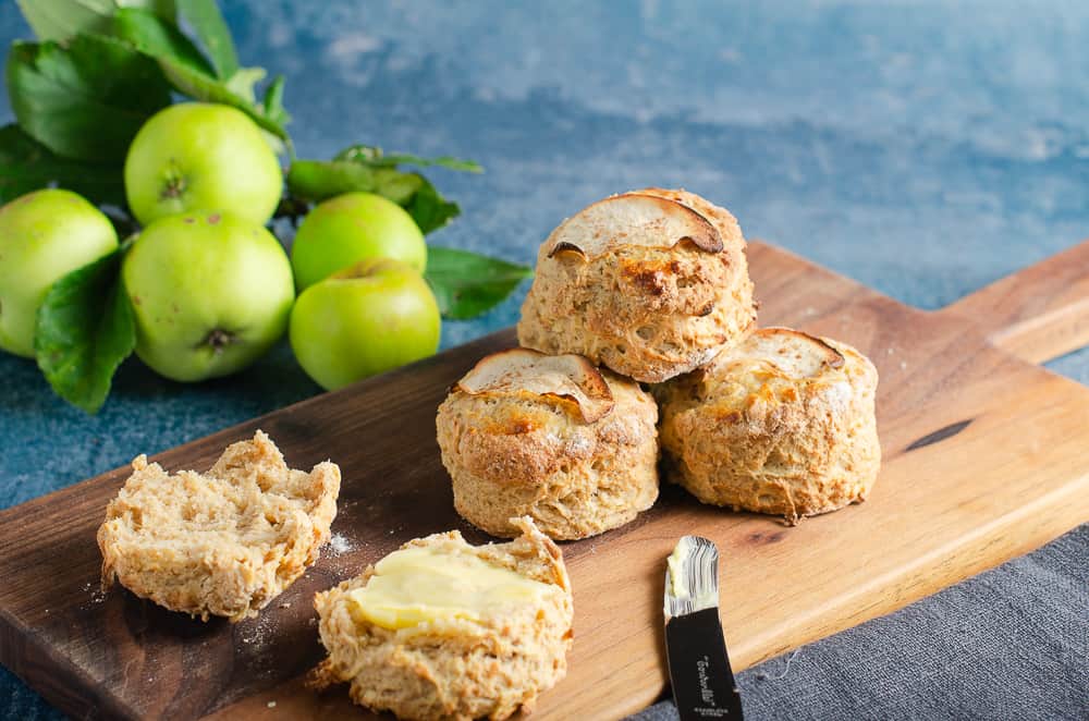 Fresh from the oven apple and cinnamon scones piled on a wooden board with freshly picked green apples still with their leaves on just slightly blurred in the background and one scone cut and ready to eat with butter on top.
