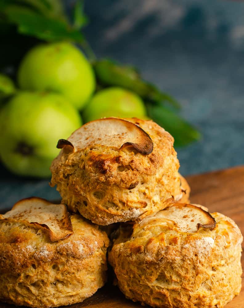 3 fresh from the oven apple and cinnamon scones piled on a wooden board with freshly picked green apples still with their leaves on just slightly blurred in the background shot close up.