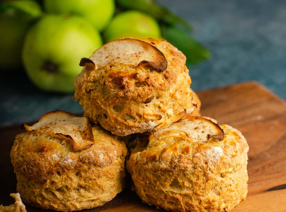 Freshly baked apple and cinnamon scones topped with a slice of apple all placed on a wooden chopping board and blue background and with freshly picked green apples from a tree still with their leaves attached.